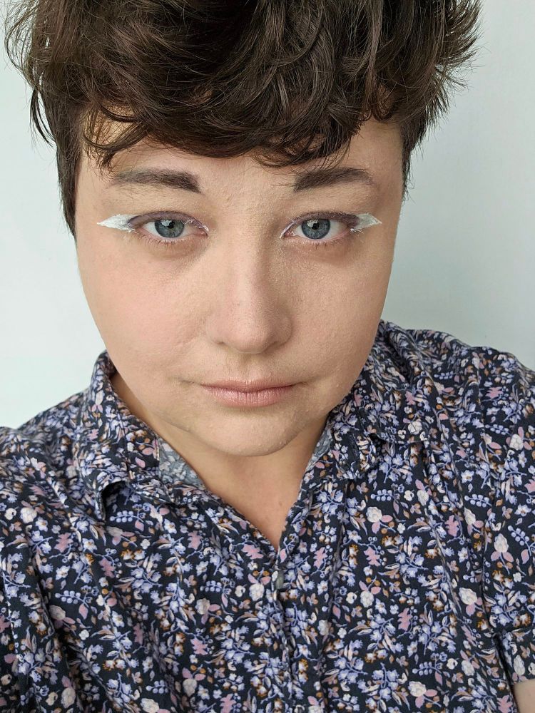 Pictured is Alejandro a white trans masc individual looking above toward the camera. They are wearing a purple floral patterned button up. Their makeup is styled with dark hazy eyeshadow and bright white eyeliner.