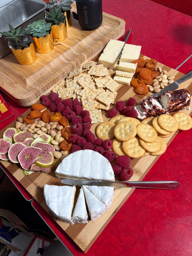 a cheese board filled with a brie, a goat cheese, and a cheddar cheese. the board is filled with crackers, almonds, dried figs, dried apricots, and raspberries 