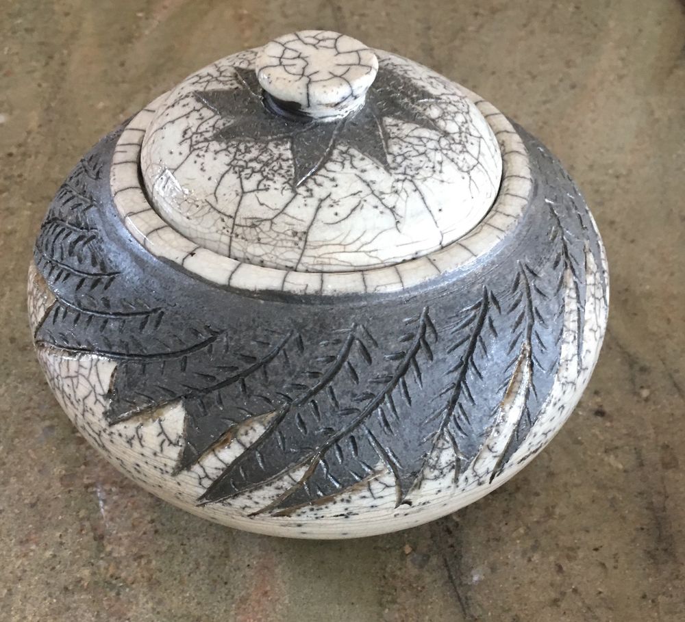 6 inch diameter bulbous jar with lid. White crackle raku glaze with carved black unglazed leaf design on lid and jar.