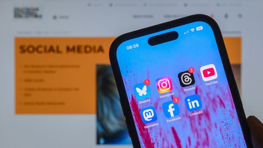 Smartphone mit den Apps von Bluesky, Instagram, Threads, Youtube, Mastodon, Facebook und LinkedIn vor dem Hintergrund der unscharfen Seite zu den Social Media der DNB