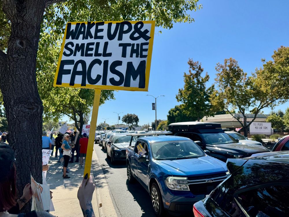 Protestor holding a sign that reads “Wake up & smell the fascism.”
