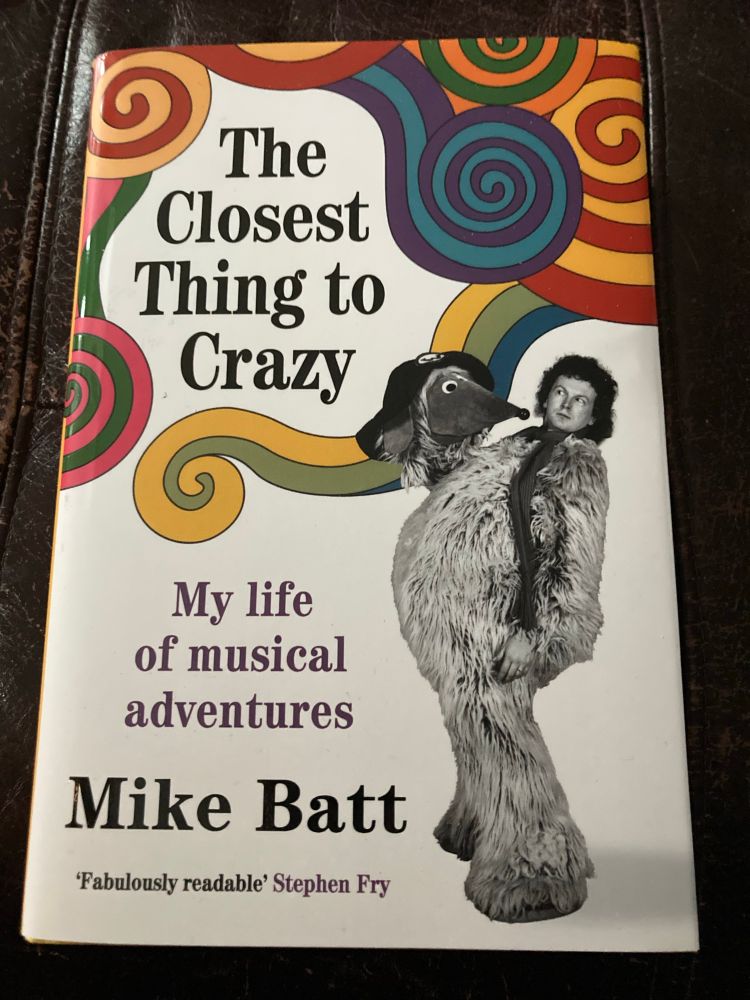 Front cover of Mike Batt’s Autobigraphy - The Closest Thing to Crazy - My life of musical adventures.
Colourful Swirls with a Black and White photo of Mike in an Orinoco Womble Costume - side profile holding Orinoco’s Head as he has looks at it with curiosity 