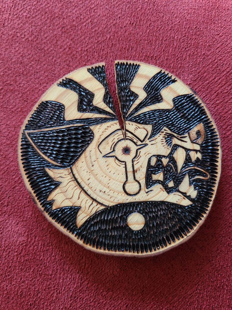 Small, round wood slice, with a large crack in the top. There is a woodburning illustration of an upset canid, smarling and yelling as the crack pierces their head. They are crying, and there are accenting zigzag lines coming from where the crack meets the scalp; to signify a splitting headache.