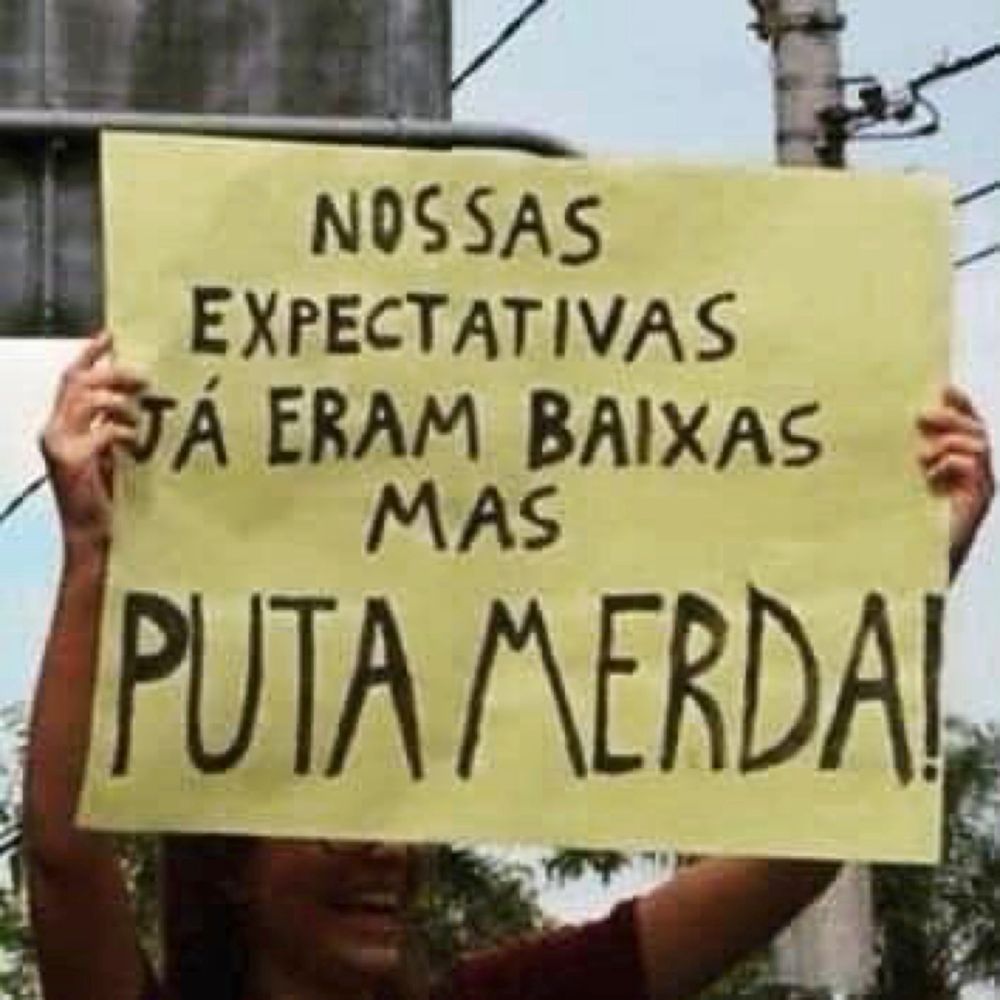 Pessoa levantando cartaz escrito NOSSAS EXPECTATIVAS JÁ ERAM BAIXAS MAS PUTA MERDA!