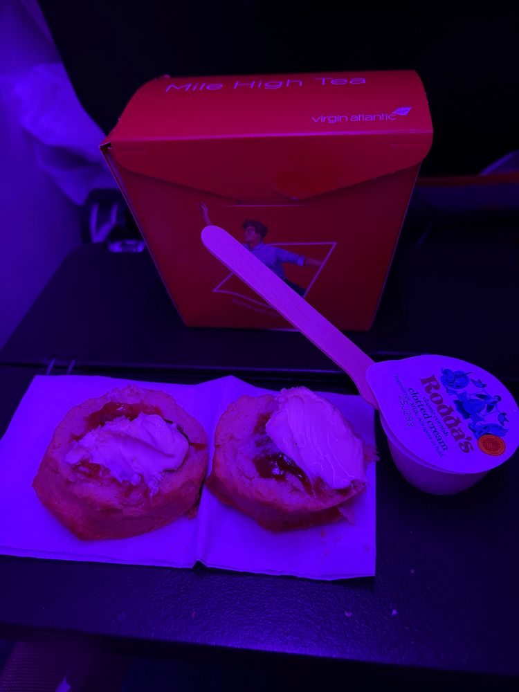 A scone, sliced in half on a serviette. Each half has a red jam on with clotted cream on top. To the right is a small pot of Rodda’s clotted cream with a wooden knife sticking out. Behind is a red cardboard box. All the items are on an airplane seat tray. 