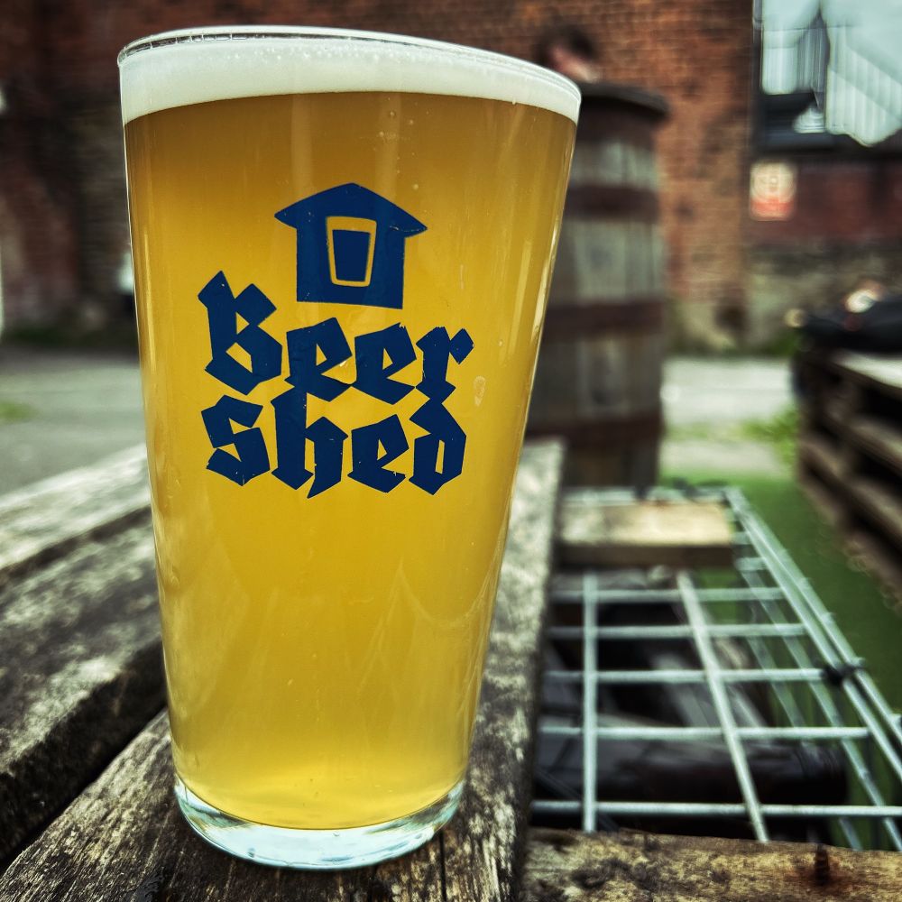 Picture of a pint of beer on a wooden table. The pint glass has the name of the pub on it “Beer Shed”.