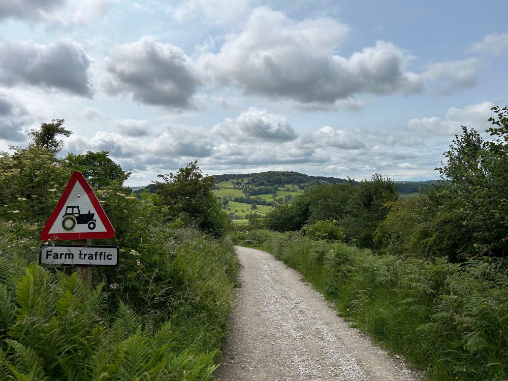 Gravel bridleway trail looking down to hills in the distance. A sign on the left shows a tractor & the words Farm Traffic.
