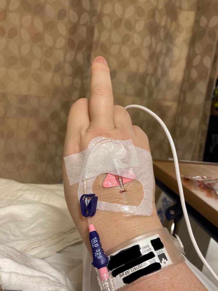 A persons hand with a pink and blue IV inserted into the back side of their hand. With the white gauze and the white printed script on the IV it makes up the colors of the transgender pride flag. Also the hand is flipping the middle finger to all the shitty bigots who told this person they would regret top surgery and it was mutilation. They don’t regret it and it’s gender affirming care not mutilation. The background is clearly a hospital setting showing this person in a hospital bed with an ugly tan privacy curtain drawn. 