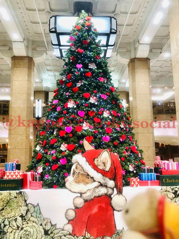 日本橋タカシマヤの1F、ヒグチユウコさんデザインの猫サンタ横顔のプレートの後ろには
大きなクリスマスツリーに赤とピンクのハートと猫オーナメント、それらを見つめるピンぼけシルバニア赤ちゃん（シルクネコ）ぬい撮り写真です