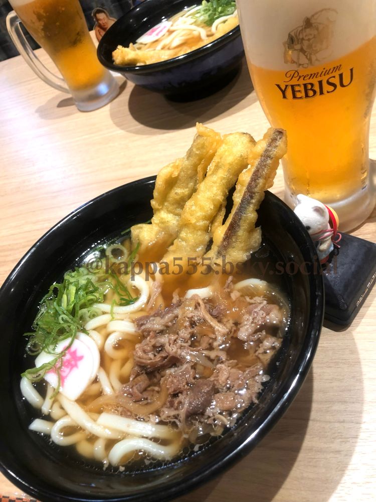 「資さんうどん」にて、柔らかく味付けしたら肉と「資」マークのかまぼことネギとスティックみたいなゴボウ天ぷらが3本のってるうどんとエビスビールのジョッキに挟まれてるシルバニア赤ちゃん（シルクネコ）ぬい撮り写真です