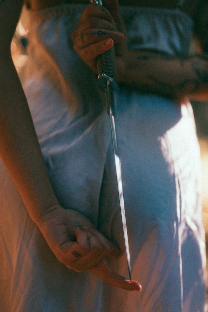 A close up of Mari holding a dagger behind her back. The point is rested on their finger and shining in the sun. 