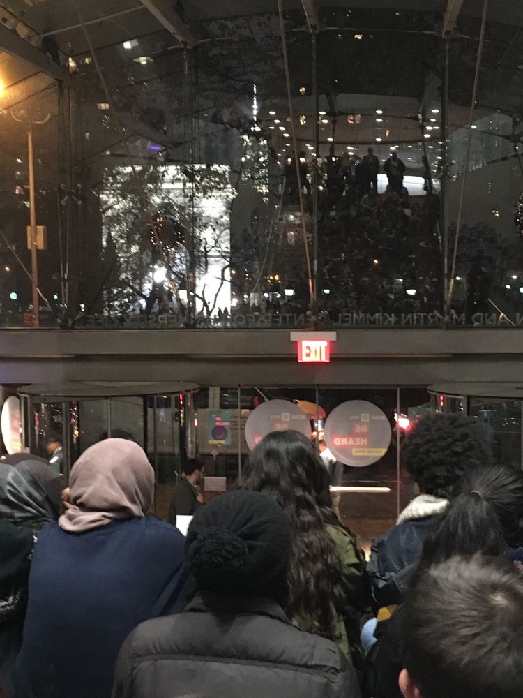 of heads and backs of people standing in a crowd staring out large window at night scene. Washington Square arch visible through window.