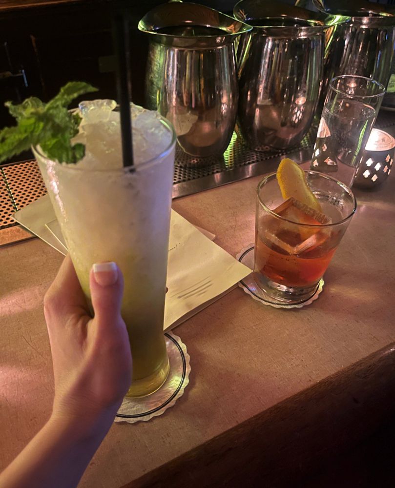 Shot of two drinks on the bar top at an establishment. On the left in the foreground is a tall greenish swizzle cocktail with crushed ice and a mint sprig. Naughty Elena is gripping said tall glass with her left French manicured hand, similar to how she would grip something else…on the right is her date’s drink, an old fashioned with one big rock in it. Two paper menus are on the bar between the glasses. In the background are frosty silver pitchers of water. Damn do I love a tropical cocktail on summer nights. 🌴🍍