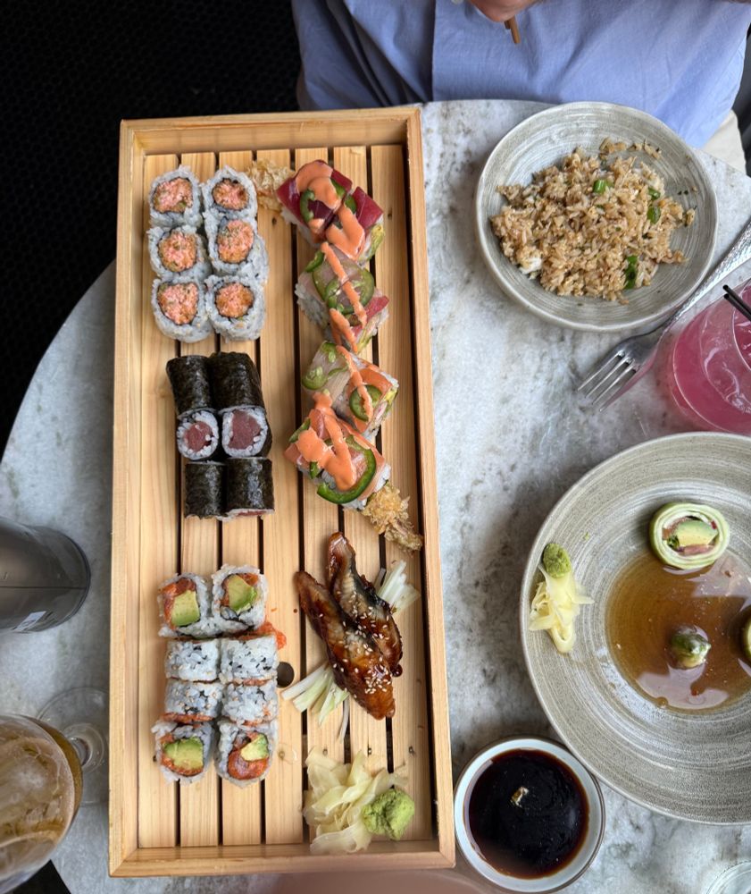 Shot from above of the table between Elena and her date at a sushi-focused restaurant. The round white marble table has a wooden tray of a variety of sushi, a small bowl of brown fried rice, drinks for Elena and her date, and a mostly-empty bowl of brussels sprouts (absolutely delicious). You can also see the fuzzy outline of Elena's thumb in the bottom of the frame because she wasn't paying attention lol.