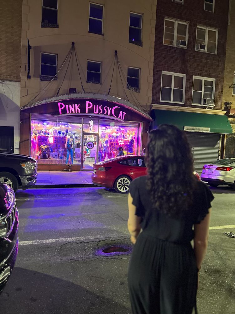 Elena is standing on the sidewalk across the street from a store for dancewear in Philadelphia called "Pink Pussycat". SHE felt like a pink pussycat so obviously she had to stop. She is facing away from the camera off to the right of the store. She's wearing a black linen romper with short ruffled sleeves. Her hair is somewhat curly and loose, falling down her back. Her bum is covered but you can see it's plump and grabbable through the thin fabric. In the background there's a red Prius in front of the store kind of throwing off the shot but whatever, the pink neon lights and signage for the Pink Pussycat is still 10/10.