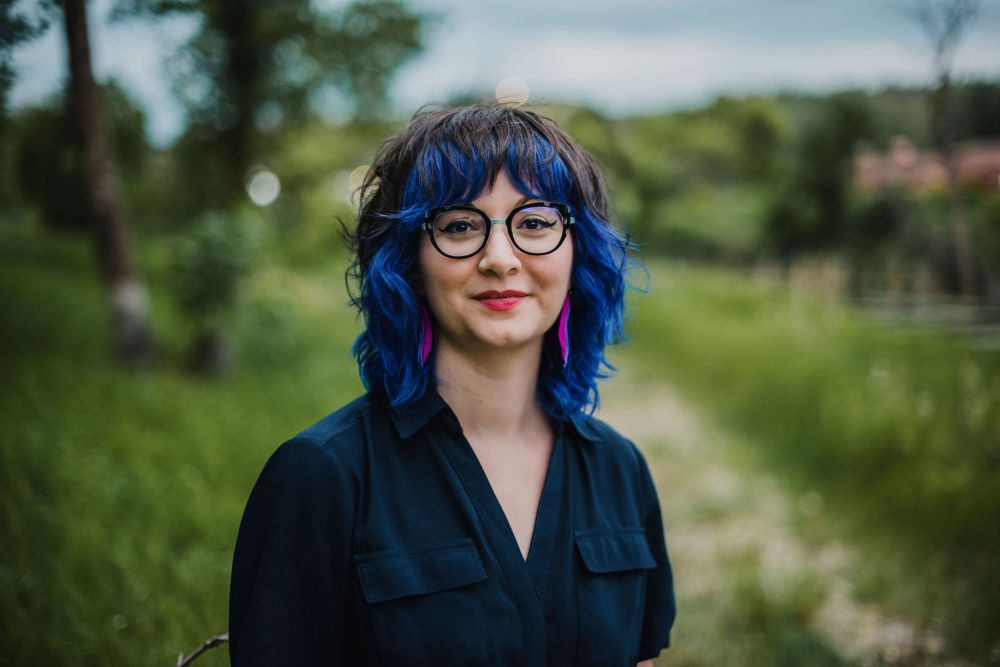 Headshot of Courtney Bates-Hardy, outside in front of a deliberately-blurred background of green grass, a few trees, a path, and some blue sky with clouds