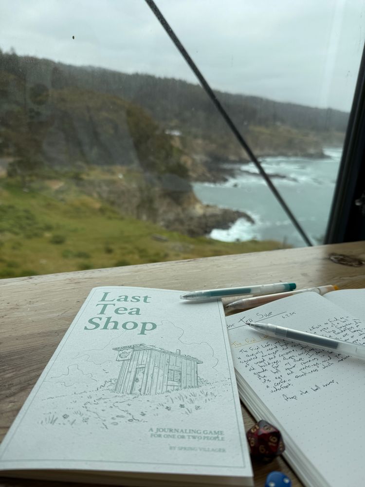 An RPG Zine titled Last Tea Shop with a tea stall drawing on the front. Adjacent is an open journal with some writings, words, and pens. The backdrop is the inlet cove of the Pacific Ocean. Overcast with the coastline in the background. 