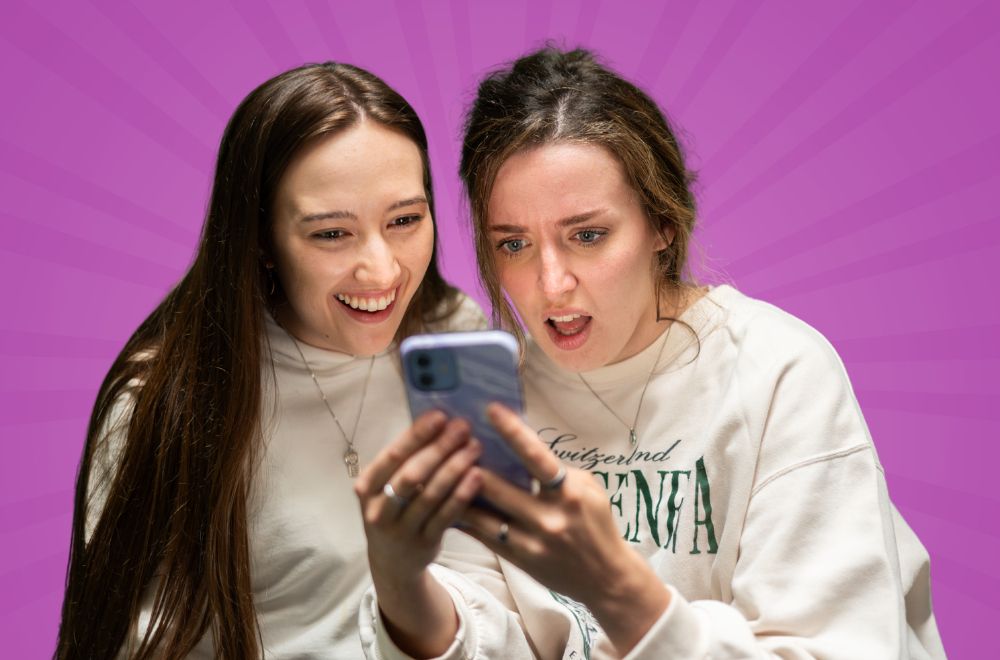 Two young woman looking at a phone screen. One is smiling, the other looks a little shocked.