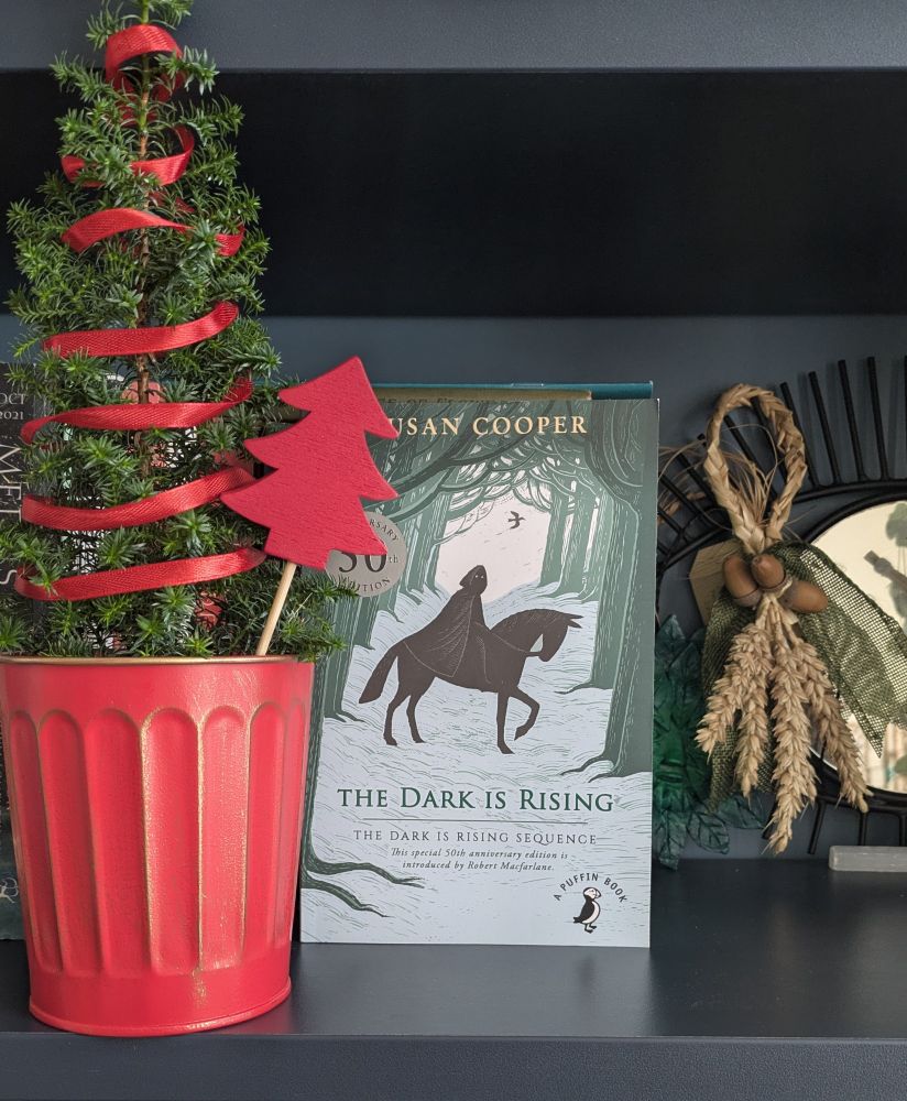 A copy of Susan Cooper's classic children's novel, THE DARK IS RISING. The cover shows a shadowy green woodland in the snow, indicating the tale happens in midwinter. In the centre of an alleyway of bare trees, a dark, hooded rider sits astride a black horse. The rider stares out of the book at the reader, as if noticing them watching. It creates a frightening atmosphere. This cover marks the 50th anniversary edition of the book, as confirmed by a silver embossed emblem on the upper left of the cover. To the left of the books sits a minute evergreen tree, decorated with red ribbon. To left, a green man mask can be seen peeping out, a corn dolly beside it