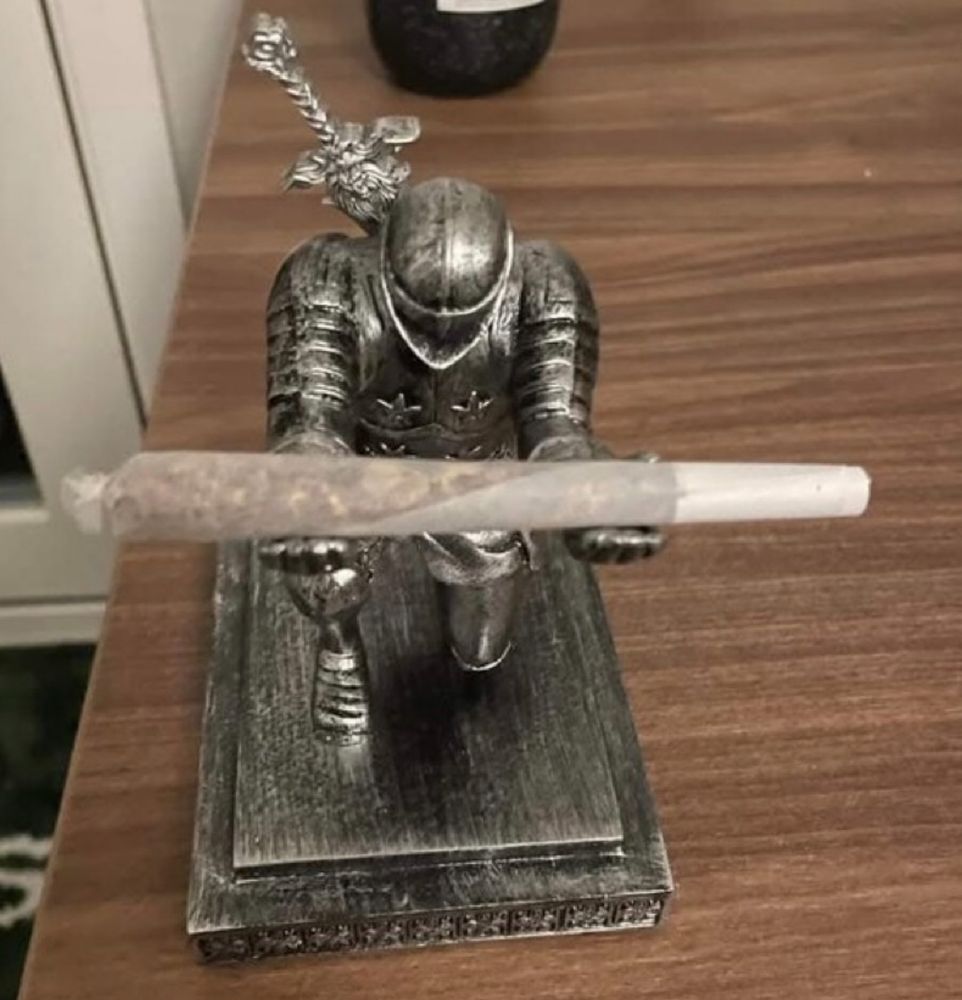 Photo of a small statue of a full dressed knight, kneeling with his head bowed, holding a beautifully wrapped joint.