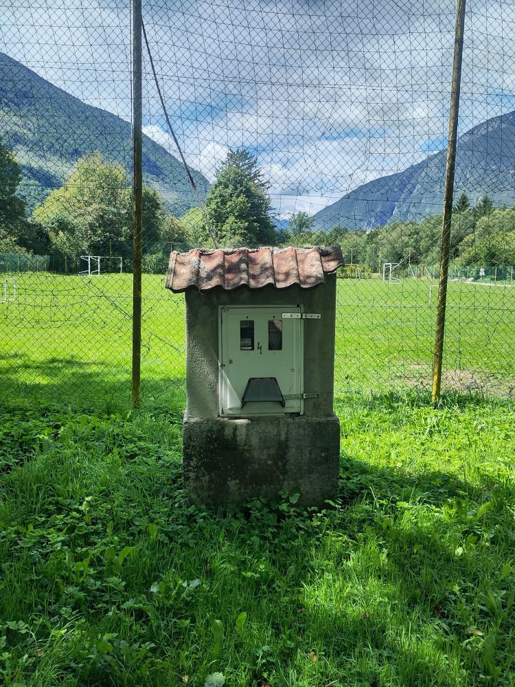 Ein Elektrokasten dessen Elemente angeordnet sind wie ein trauriges Gesicht steht vor einem Zaun eines Fußballfeldes in den Bergen.
