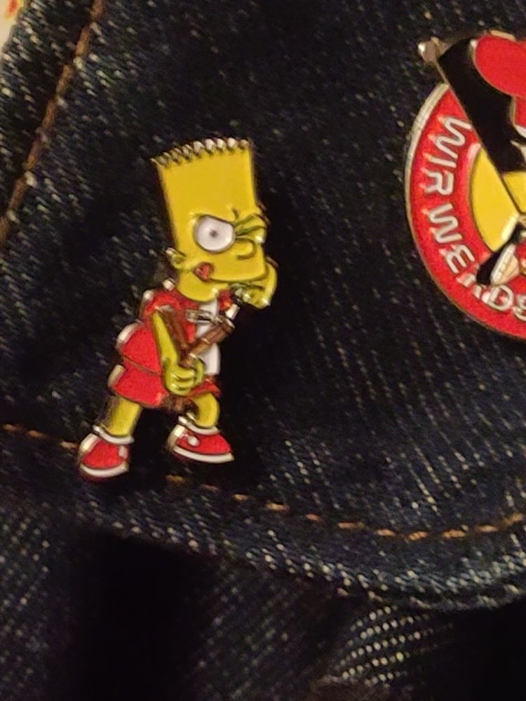 An einem Jeanskragen ist ein Bart Simpson Pin in Rot Weißem Union Trikot und einer Zwille in der Hand.