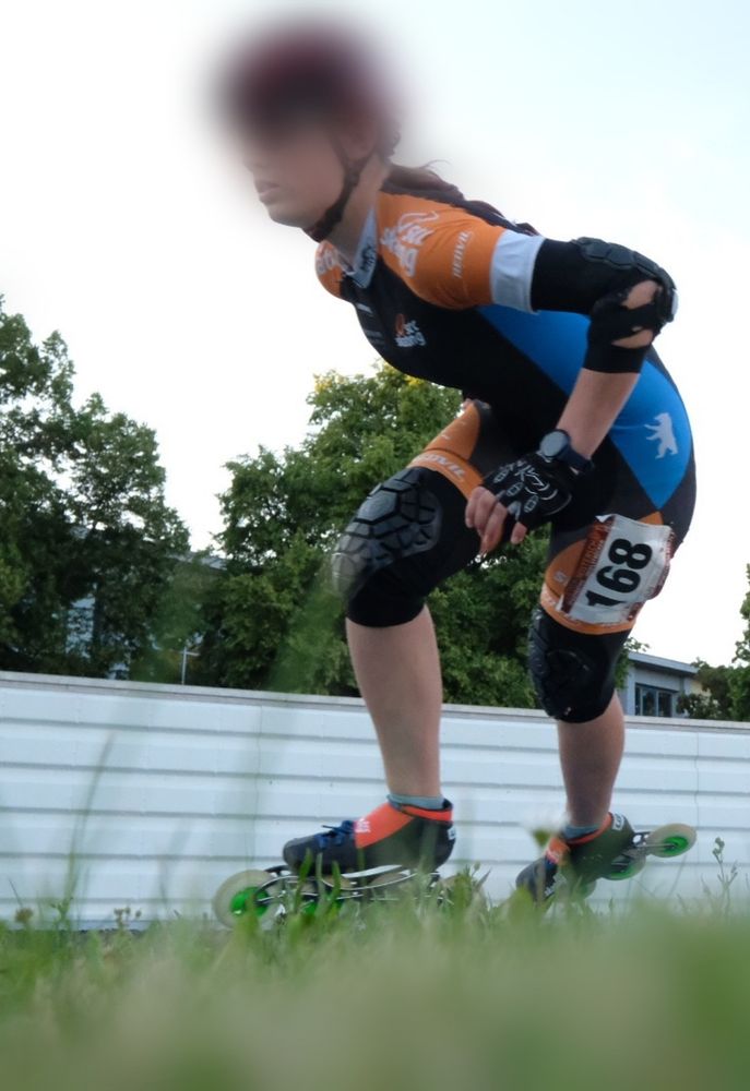 Eine Speedskaterin von unten fotografiert liegt schräg in der Kurve. Im Vordergrund Rasen im Hintergrund Bäume und Himmel.