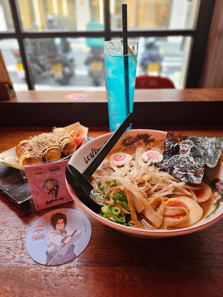 All items atop a wooden countertop table. On the right, a Yokocho x Naruto Shippuden themed bowl, featuring a dragon and text reading 'London' on the inside rim of the bowl. The provided description is as follows: Teuchi's famous 3hrs miso based chicken & pork broth topped with extra char siu pork belly, spicy umami red paste. Nitamago egg, menma, beansprouts, naruto & spring onion. All finished with Naruto nori and served in our limited edition Yokocho x Naruto bowl!

To the left, a collaboration coaster with the character Sasuke Uchiha as the design, with a small enamel pin badge of Sakura Haruno behind it in her Shippuden outfit (red jacket and shorts) A portion of Takoyaki (6 pieces) is to the back corner - fish flakes, mayo and red pickles are on top, with sauce dripped over it. At the back centre, the Sasuke Uchiha Soda in a slim, tall glass. Light blue in colour and containing a dried lemon at the top of the glass.

In the background, a window facing the road/pavement, with a row of bikes neatly parked.

