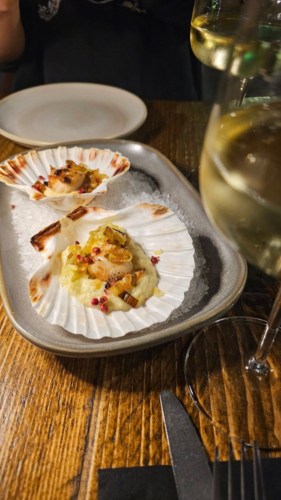 Pictured: Scallops served on a bed of ice and a metal tray. To the right, a pair of wine glasses filled 1/4 with white wine. A knife partially appears at the bottom of the screen