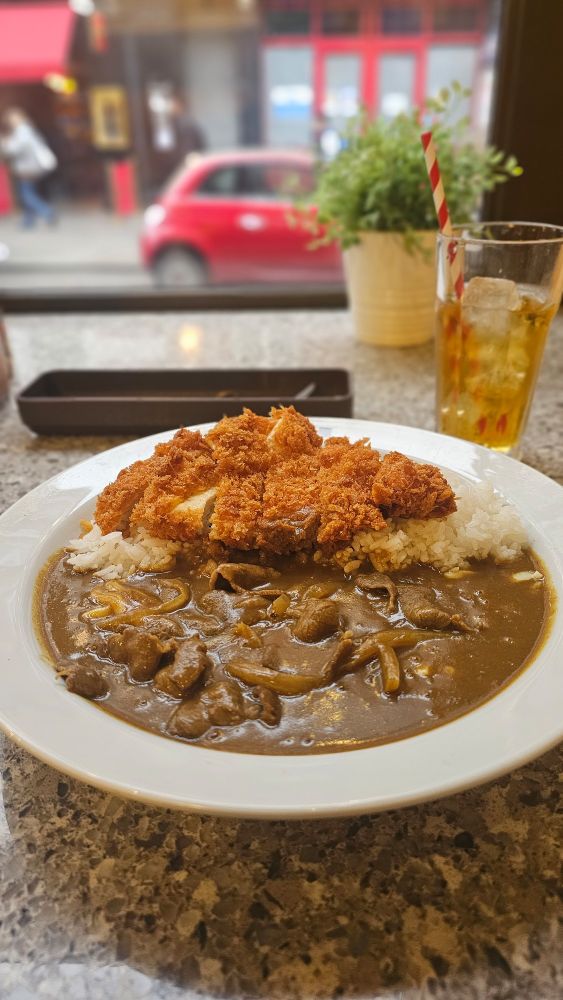 All items placed on top of a marble counter top. Front and centre, a plate branded with the coco ichibanya logo filled with curry sauce, thinly sliced beef, and cheese, further back chicken katsu ontop a bed of rice - 350g. In the back right, a glass of apple juice with a red and white straw. A fake plant is at the right corner of the table, with a cutlery tray on the far left. A window is visible in the background with a red car parked across the road.
