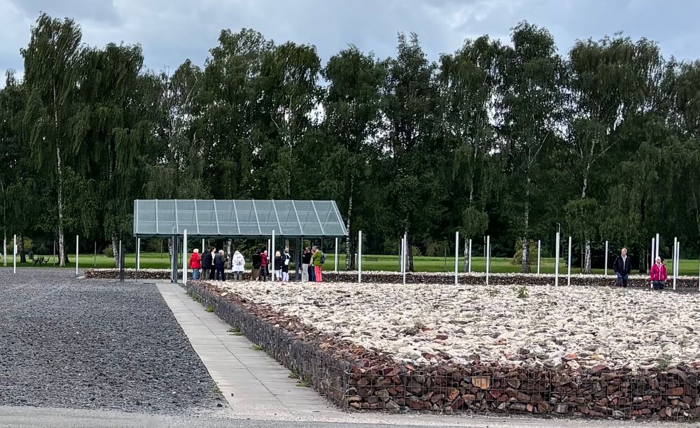 Blick auf Steinhaufen im Vordergrund, Bäume im Hintergrund.  Dazwischen eine Gruppe Menschen, die unterbringen Glasdach stehen 