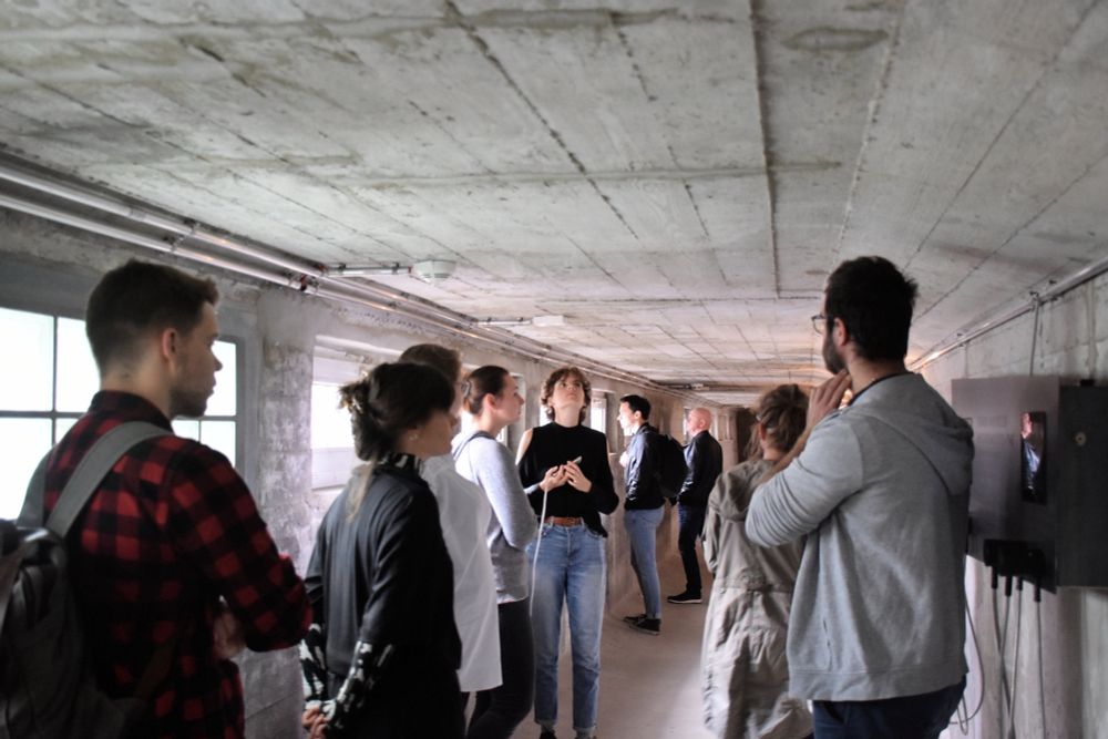 Eine Personengruppe steht in einem Gang mit niedriger Decke. Die Wände sind aus Beton. 