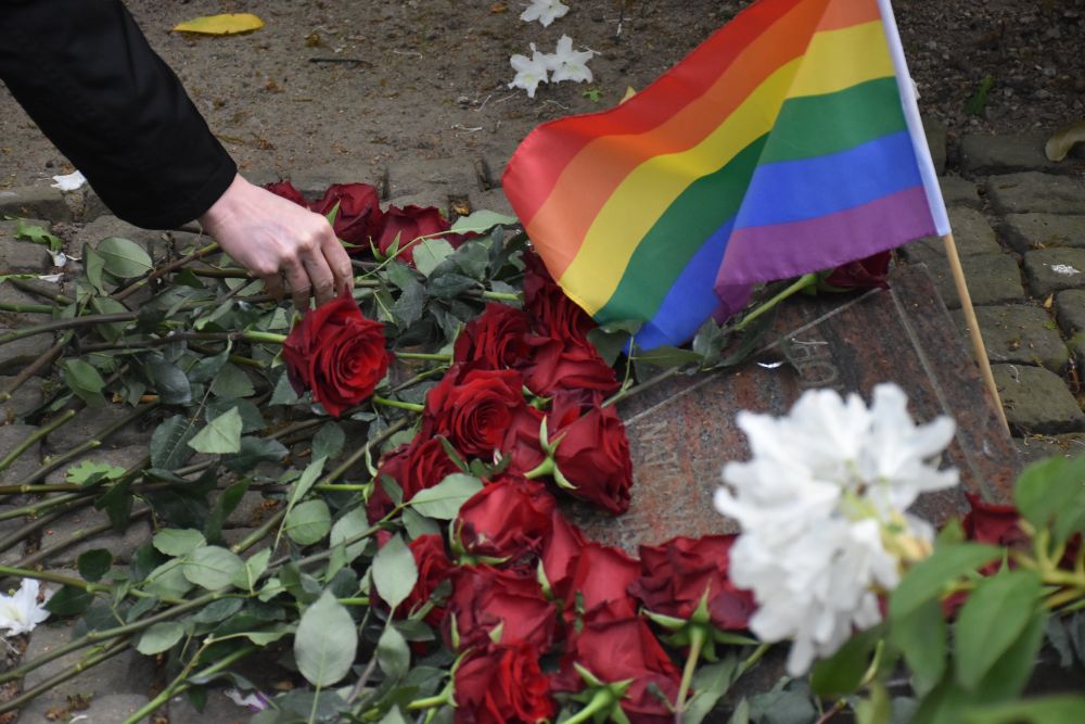 Eine Regenbogenfahne und viele rote Rosen liegen auf einem Gedenkstein, der an homosexuelle Häftlinge erinnert. Eine Hand legt eine weitere Rose dzu.
