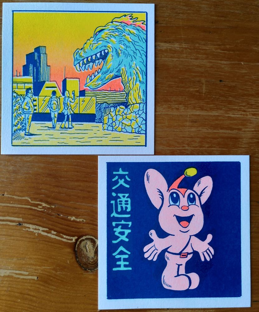Photos de deux cartes postales dessinées par Simon Bournel-Bosson. L'une représente une statue d'une tête de Kaiju au milieu de Tokyo et prise en photo par des badauds. L'autre représente un petit personnage kawaï qu'on pourrait assimilé à une souris anthropomorphe rose, sur fond bleu marine.
