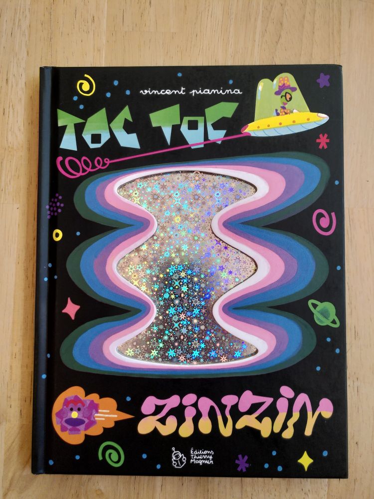 Couverture de l'album jeunesse Toc Toc Zinzin de Vincent Pianina. On y voit au centre une forme non-identifiée, brillante avec des étoiles incrustées, le tout sur fond noir. Au-dessus, il y a une soucoupe volante avec une OVNI au volant et autour des formes nous faisant penser à l'espace.