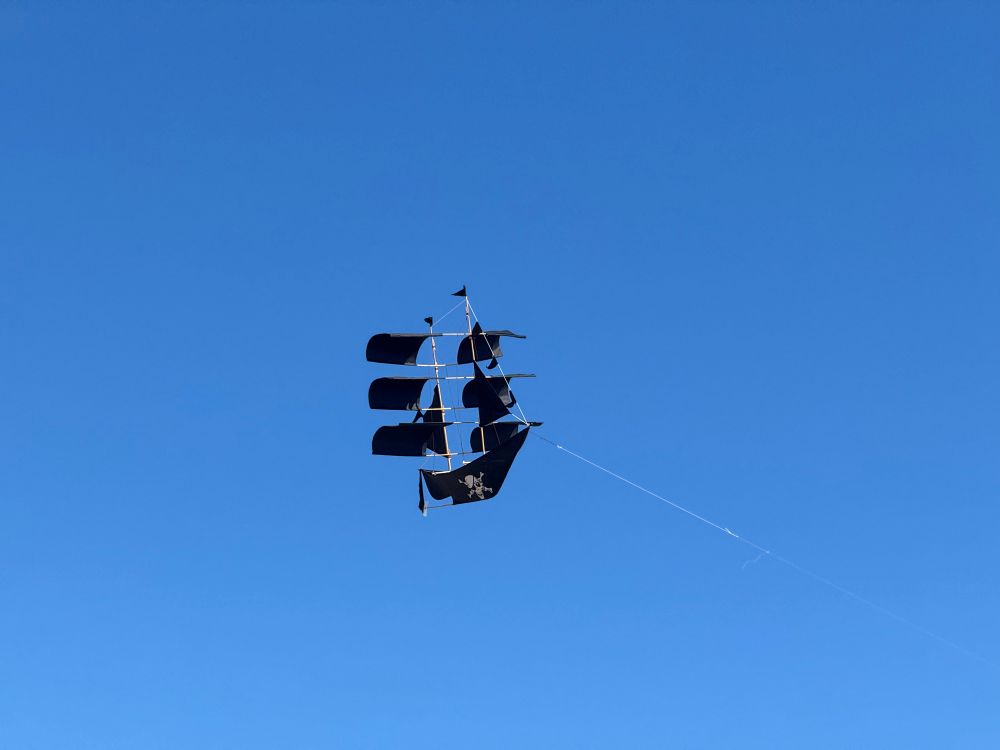 A black kite against a blue sky made in the shape of a pirate ship, and apparently flying, much to the disbelieve of the off camera owner and his kids!