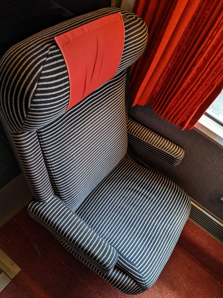 SNCF 1st class Corail coach showing a single seat. The seat has fine black and white stripes with a red antimacassar. Red curtains are seen on the edge of the photo.