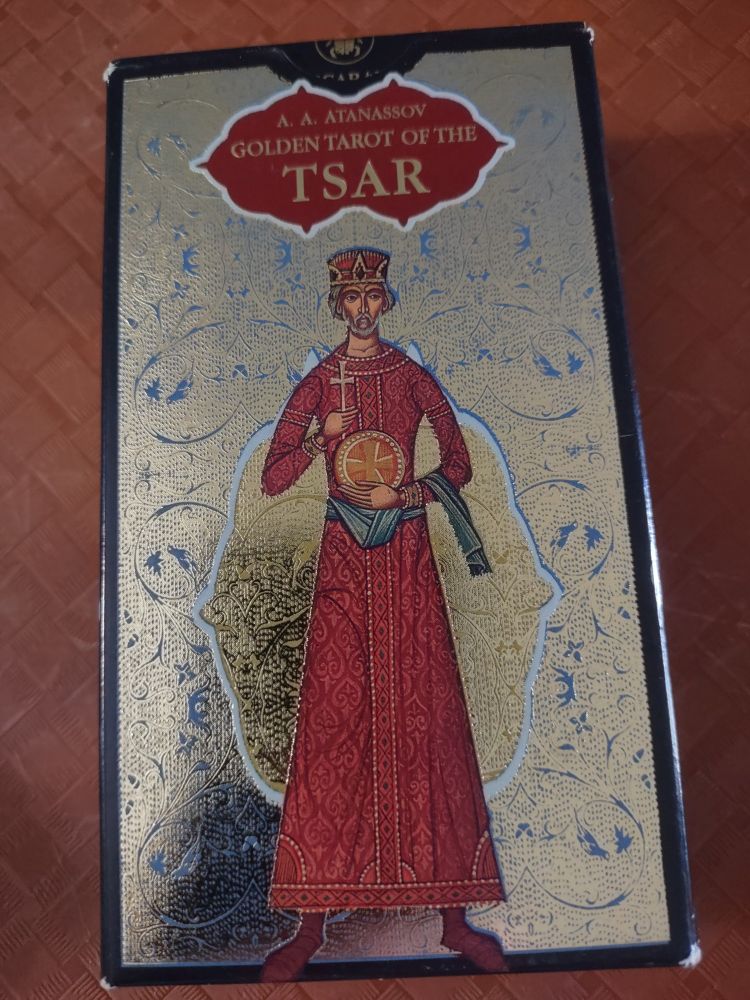 Deck box with a background of navy blue that borders all sides. Next layer is gold foil with pale blue leaf design. At the top is a fancy ovalish shape (like two acorns head to head). It's gold lettering upon an orangey red background bordered by white. Underneath that is a man upon a gold design with white border that resembles a cupcake with icing shape. The man has a red hat with a crown design. He has short gray hair; a gray mustache and a short gray beard. He is wearing an orangey red dress and shoes. The dress has golden stitching with a faded spade design. There is a narrow blue scarf wrapped around his waist. He is holding a cross in one hand. The other hand holds a gold plate with a wide gold cross on it.