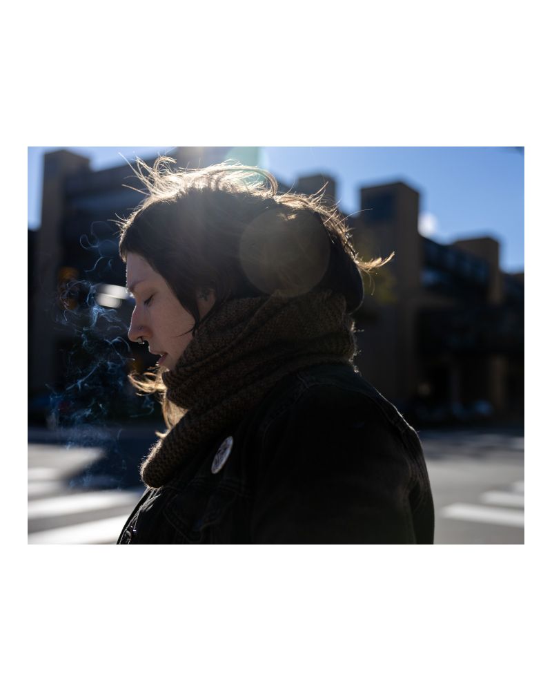 A person wearing a scarf stands in profile outside, backlit by the sun. Wisps of cigarette swirl in front of their face. 