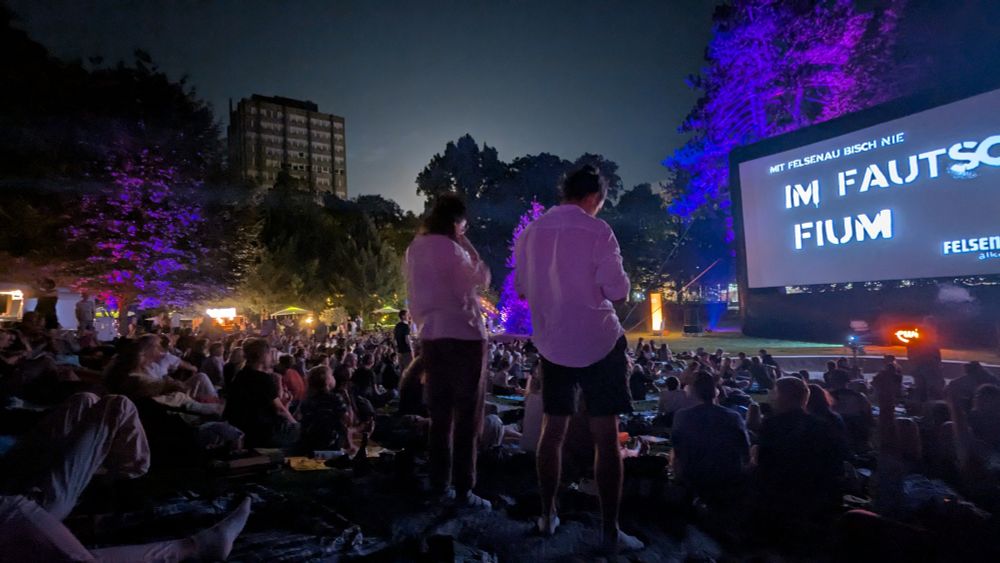 Open-Air-Kino: man sieht die Leinwand, es ist finszer und zwei Zuschauer:innen stehen zwischen vielen Zuschauenden. Rechts ist die Leinwand sichtbar. 