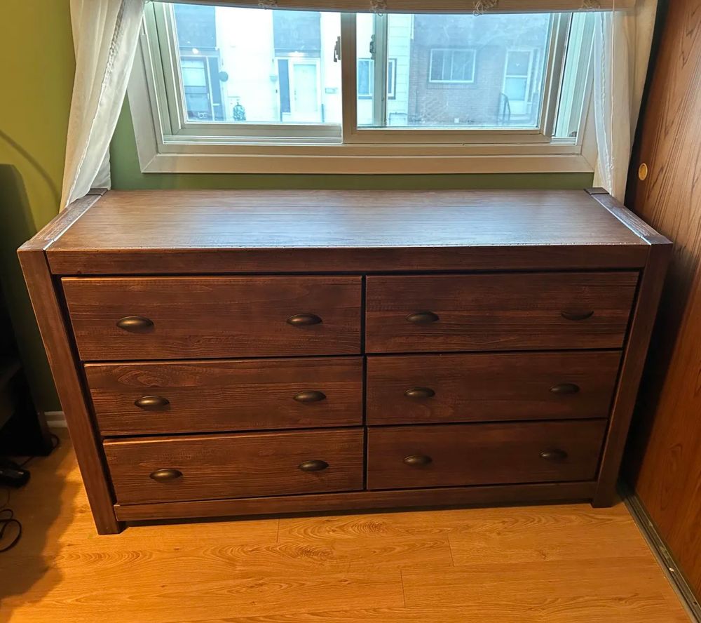 A brown, wooden set of dresser drawers, two wide and three high, sitting under a window looking outside.  The floor's a kind of orangey wood, and the wall in the back is a sage green.
