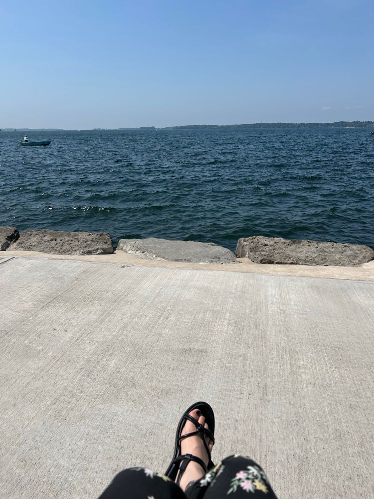 Feet in sandals crossed on a stone walkway. Water and blue sky in the background. 