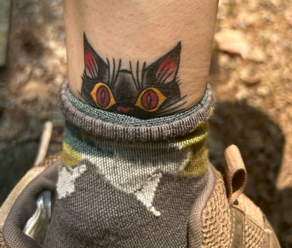A black cat tattoo in the American traditional style peeks out from a hiking sock. 
