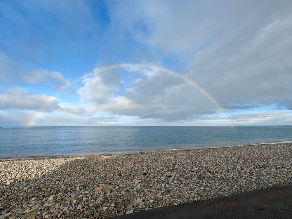 A long shot of a pebble beach and the calm sea beyond. The sky is blue with white clouds, and the full arch of a rainbow can be seen over the sea. 