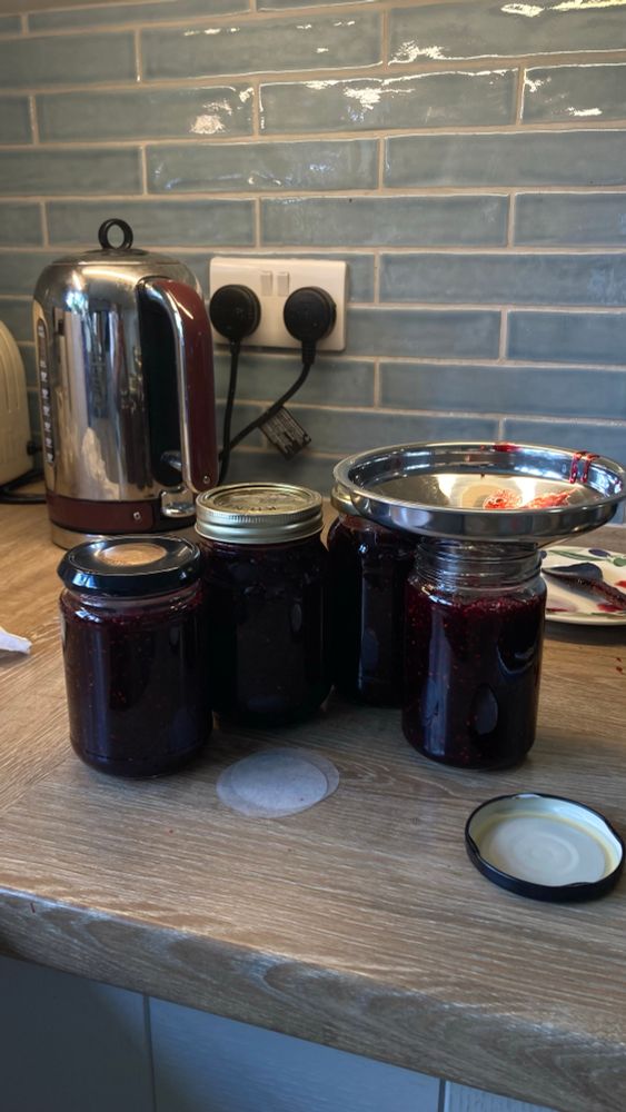 Four jars of homemade jam on a kitchen counter. The jar in the right has a jam funnel on top of it. 