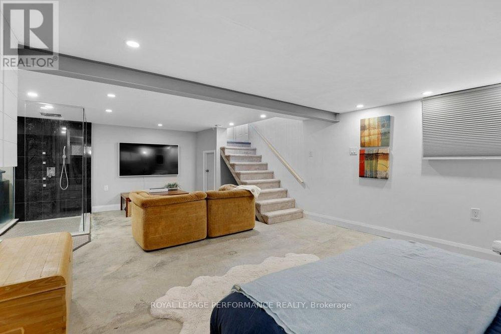 realtor picture of a basement suite looking out from the bed. beige carpet. tan furniture and large television on the far wall. glass walled shower fully visible to the rest of the room.
