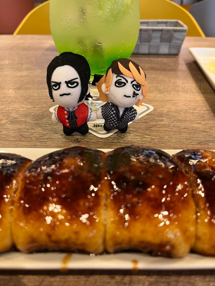 焼きまんじゅうを暖かく見守る櫻井敦司と今井寿のぬい。焼きまんじゅうは蒸しパンのようなふわふわ食感に甘い味噌だれをかけて焼いたものです。