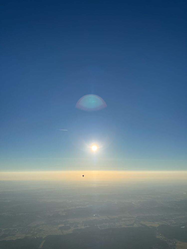 Blick aus dem Korb eines Heißluftballons in 1.500 Meter Höhe. Die Sonne ist mittig des Bildes und etwas über dem Horizont. Über der Sonne spiegelt sich ein Halo. Der Himmel ist blau. Die flache Landschaft ist leicht in Dunst gehüllt. In weiter Ferne unter dem Horizont ist ein Heißluftballon.  