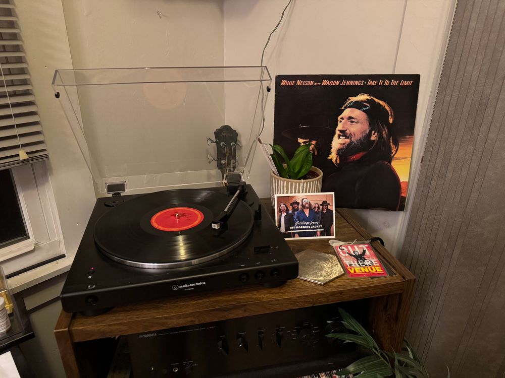 A turntable with Willie Nelson & Waylon Jennings’ album Take It to the Limit on the platter. Next to it, the album sleeve, a potted plant, a My Morning Jacket postcard, a gold coaster, and a lanyard from Paul McCartney’s Out There Tour. 