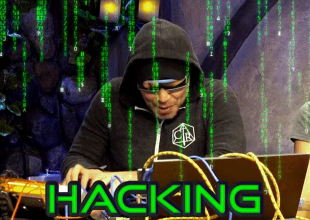 a man in a hoodie is using a laptop and the word hacking is on the screen behind him